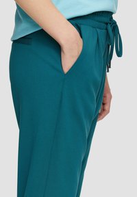 Tealowe spodnie dresowe z elastycznym pasem, miękka tkanina, boczne kieszenie oraz plisowane detale z przodu, które dodają stylu i komfortu.