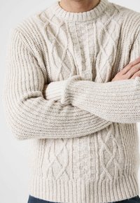 Mann trägt cremefarbenen Zopfmuster-Pullover, Arme verschränkt, sichtbar vom Hals bis zur Hüfte vor weißem Hintergrund.