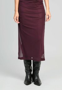 Femme portant une jupe bordeaux transparente midi associée à des bottes en cuir noires montantes, se tenant contre un fond uni.