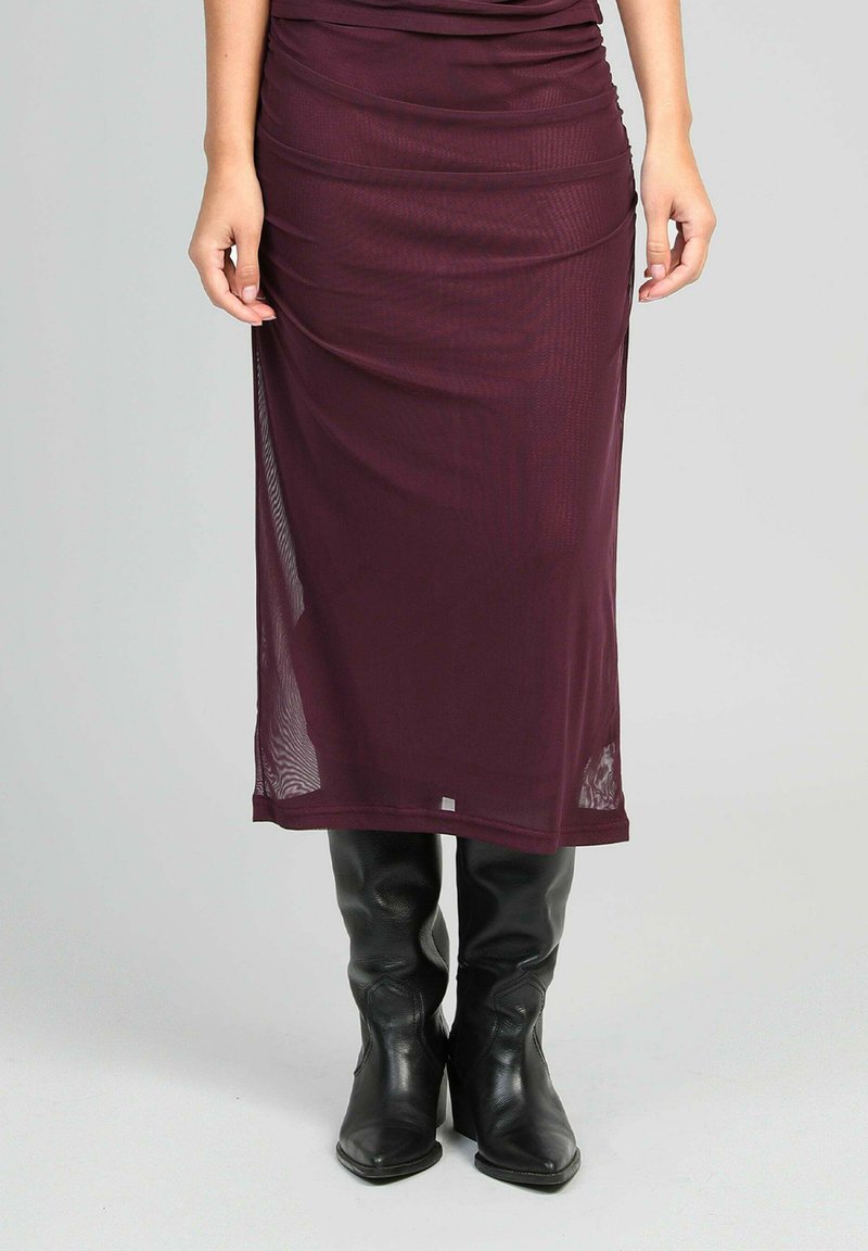 Femme portant une jupe bordeaux transparente midi associée à des bottes en cuir noires montantes, se tenant contre un fond uni.