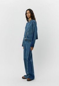 Giubbotto di jeans con un design cropped e bottoni visibili, abbinato a jeans a gamba larga. Entrambi i capi sono di un blu medio e presentano una texture morbida.