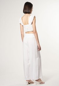 Witte cropped top met kantdetail en ruffles op de schouderbanden, gecombineerd met losse witte broek en witte sandalen met bandjes.