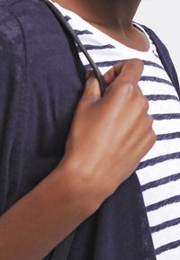Námořnický kardigan přes bílo-modře pruhované tričko. Ruka drží černý popruh. Tkanina vypadá lehká a strukturovaná.