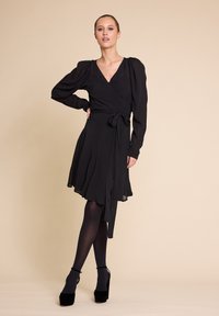 Svart omslagkjole med lange puffermer, en knyttet midje og en svevende midje. Kombinert med svarte tette strømpebukser og høye hæler.