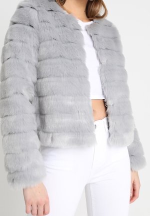 Vrouw draagt een lichtgrijze, horizontaal doorgestikte faux bontjas over een witte cropped top en witte hoge taille broek.