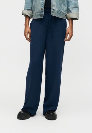 Osoba nosiaca voľné tmavomodré nohavice so sťahovacou šnúrkou, čierne masívne topánky na platforme a svetlú denimovú bundu s vyhrnutými rukávmi.