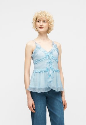 Młoda kobieta z krótkimi, kręconymi blond włosami, ubrana w bezrękawową, jasnoniebieską bluzkę z falbanami i niebieskie dżinsy, patrząca na bok.