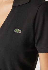 Černé polo tričko s texturovanou tkaninou. Má klasický límec a malé zelené logo krokodýla vyšívané na levé straně.