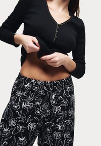 Černý top s dlouhými rukávy, výstřihem do V a detaily s knoflíky, spárovaný s černými kalhotami s květinovým vzorem a bílými konturami, které mají elastický pas.