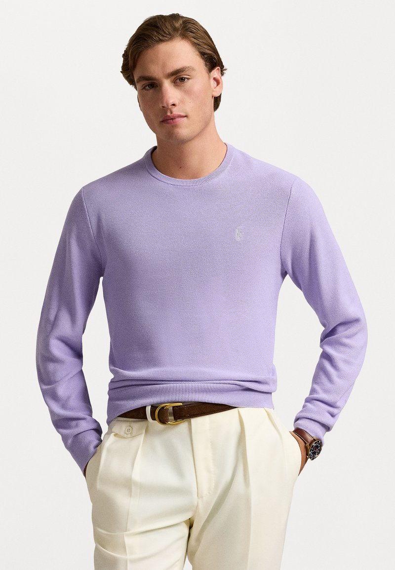 Lyse lilla sweater med rund halsudskæring og lange ærmer, lavet af tekstureret strikstof. Båret over beige bukser med et brunt bælte.