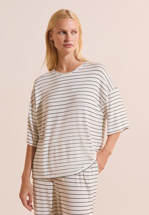 Mujer rubia que lleva una camiseta blanca de manga corta y suelta y pantalones con finas rayas horizontales negras, mirando de lado con la mano en el bolsillo.