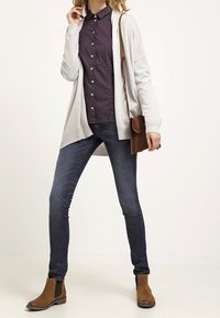 Cardigan gris clair sur une chemise à pois noire, associé à un jean foncé et des bottines brunes ; un sac bandoulière marron complète la tenue.