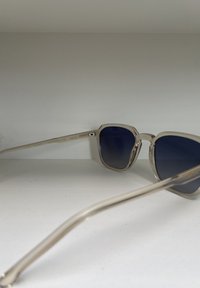 Sunglassar med en ljusfärgad metallram, mörkblå linser och en unik vinkelräffad form; har en logotyp och modellnummer på sidan.