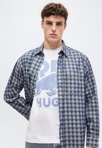 Νέος άνδρας με κοντά καστανά μαλλιά, που φοράει μπλε καρό πουκάμισο πάνω από λευκό γραφικό μπλουζάκι με εικόνα βουβάλου και κείμενο.