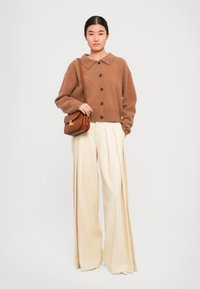 Brun strikket cardigan med skjortekrage og knaplukning, parret med lyse kremfargede vidde bukser med folder. Liten brun skulderveske.