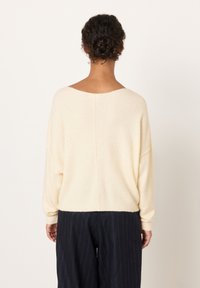 Sieviete ar tumšiem cirtainiem matiem zemā sasietā bulciņā valkā krēmkrāsas adītu džemperi un tumšas svītrainas bikses, skatoties prom pret vienkāršu fonu.