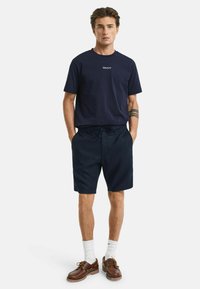 Marineblauwe katoenen T-shirt met wit "GANT" logo, gecombineerd met donkere shorts. Model draagt bruine leren schoenen en witte sokken, staande tegen een witte achtergrond.
