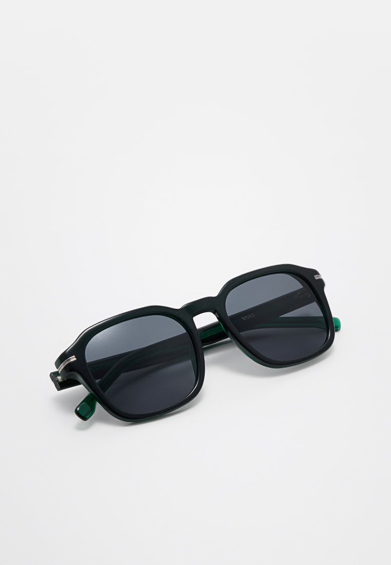 Lunettes de soleil carrées avec des verres teintés, monture en plastique vert et accents métalliques sur les charnières. Caractéristiques d'embouts d'oreilles texturés pour le confort.