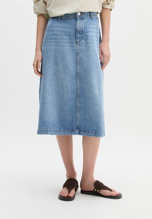 Lichtblauwe denim midirok gedragen met een beige shirt en bruine sandalen, getoond van taille tot voeten tegen een witte achtergrond.