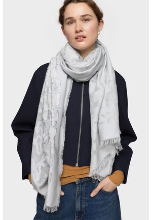 Joven con pañuelo claro con estampado alrededor del cuello, chaqueta azul marino con cremallera frontal, camiseta mostaza y vaqueros azules, manos en los bolsillos.