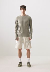 Langærmet, olivengrøn atletisk skjorte med tætsiddende manchetter parret med løse, lyse beige shorts med sidesømme. Footwear er dæmpet grøn.