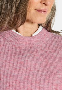 Eine Frau mit langen Haaren trägt einen gerippten pinken Pullover über einem weißen Hemd und lächelt sanft mit natürlichem Make-up und glänzenden Lippen.