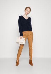 Pull V en laine marine avec une texture douce, pantalon beige ajusté et bottines à talons brillantes marron. Tenant un sac à main structuré couleur crème.
