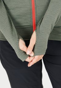 Langärmliges, olivgrünes Oberteil mit rozipper und Daumenlöchern. Leichter Stoff mit subtiler Textur. Kombiniert mit schwarzen Hosen.