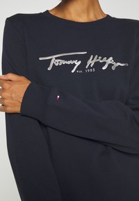 Námořnická modrá mikina s dlouhým rukávem vyrobená z bavlny, s tím, že na ní je stříbrné logo "Tommy Hilfiger" a malý trojbarevný emblém na manžetě.