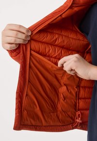 Mains tenant une veste matelassée orange ouverte, montrant sa grande poche intérieure et son ourlet à cordon de serrage réglable.