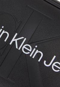 Fekete, texturált anyag kiemelkedő betűkkel. Kiemelkedő "Calvin Klein Jeans" felirat fehér színben a felületen.