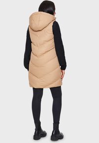 Beige wattierte Weste mit großer Kapuze und Chevron-Steppung. Getragen über einem schwarzen Langarmoberteil und kombiniert mit schwarzen Leggings und Stiefeln.