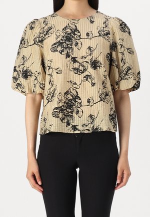 Blouse beige froissée avec imprimé floral noir et manches courtes bouffantes portée avec un pantalon noir.