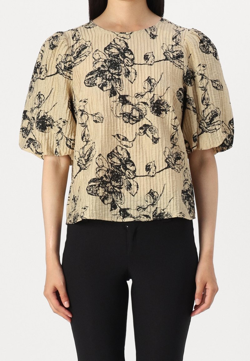 Blouse beige froissée avec imprimé floral noir et manches courtes bouffantes portée avec un pantalon noir.