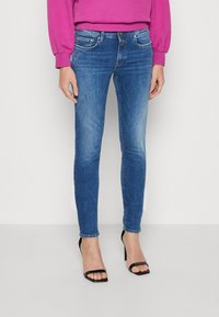 Jeans ajustados de mezclilla en color azul con un acabado desgastado. Presentan cinco bolsillos, cierre de botón y un diseño slim fit. Se llevan con tacones negros de tiras.