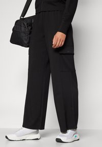 Black cargo pants feature a loose fit, side pockets, and are paired with white sneakers. The fabric appears smooth and lightweight.