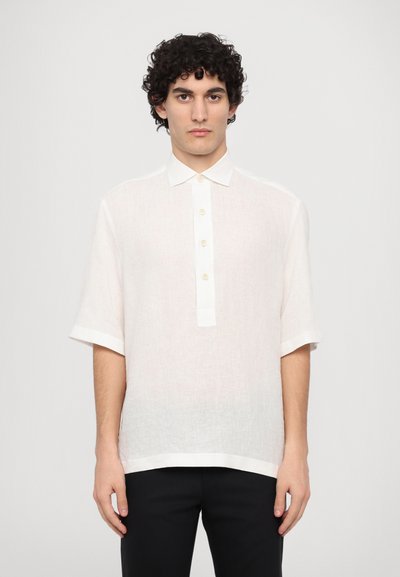 Chemise blanche en lin à manches courtes avec une patte de boutonnage, présentant une coupe droite et une texture subtile. Portée avec un pantalon noir.
