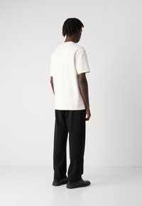 Weißes T-Shirt mit kurzen Ärmeln, kombiniert mit schwarzen Hosen. Das Shirt hat eine lockere Passform, während die Hose tailliert und gerade geschnitten ist.