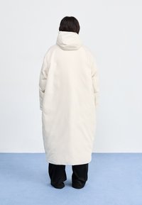 Długi, puchowy płaszcz z kapturem w kolorze off-white. Ma pikowaną fakturę, rękawy raglanowe oraz prosty krój, sięgający do połowy łydki, noszony z czarnymi spodniami.