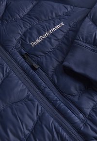 Mørkeblå quiltet jakke med frontlynlås, elastiske manchetter og "PeakPerformance" broderet på brystet. Glat, let tekstur.