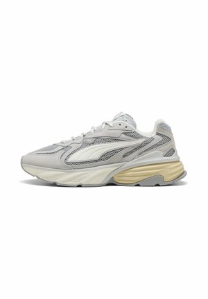 Hellgrauer und beiger Sportschuh mit Mesh- und Wildlederpaneelen, dicker profilierter Sohle und Schnürverschluss, von der Seite gezeigt.