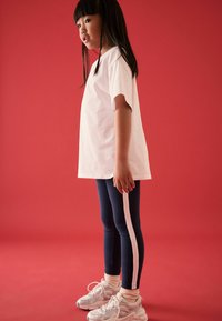 T-shirt bianco in cotone, leggings blu navy con strisce rosa ai lati e sneakers bianche con pannelli testurizzati. Sfondo rosso uniforme.
