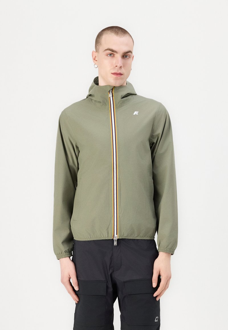 Chaqueta ligera de color verde oliva con cierre de cremallera, capucha y una raya multicolor a lo largo de la cremallera. Cuenta con puños elásticos y un logo.