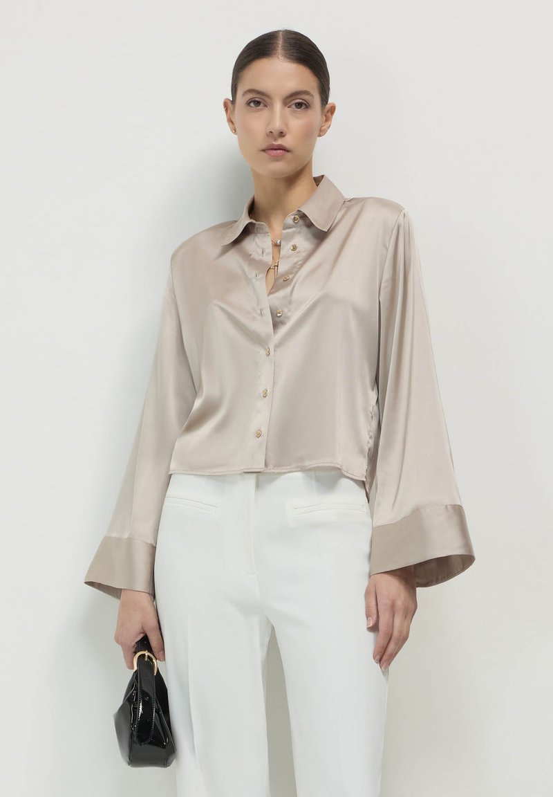 Camicia corta in seta color beige con colletto, chiusura a bottoni e maniche ampie con polsini in satin a contrasto. Il modello tiene in mano una piccola borsa nera.