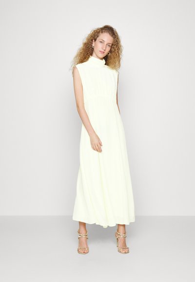 Abito maxi senza maniche di un leggero giallo con colletto alto, vita arricciata e gonna fluida; abbinato a sandali con tacco alto impreziositi da dettagli metallici.