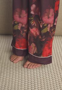 Mély bordó alapon virágmintás nadrág rózsaszín és zöld díszítéssel; kontrasztos egyszínű mandzsetta és fehér szegély; mezítelen lábak egy texturált világos szőnyegen.