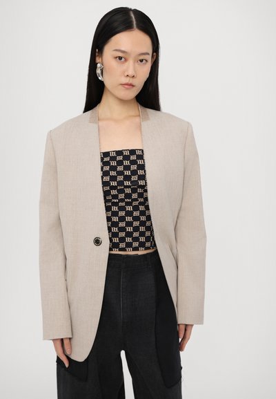 Beige linned blazer med en single sort knap, skræddersyet design og nedskårne klapper, båret over en sort mønstret top og mørke bukser.