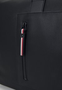 Schwarze Handtasche mit Textur, die über eine Reißverschlusstasche mit einem gestreiften Ziehgriff in Rot, Weiß und Blau verfügt, ergänzt durch glatte schwarze Akzente.