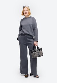Kvinde med kort blondt hår iført mørkegrå sweatshirt og bukser, sorte hælede sandaler, solbriller på hovedet, holder en grå og sort håndtaske.