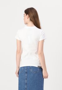Witte, T-shirt met korte mouwen en ronde hals, gedragen onder een blauwe denim rok. De stof lijkt glad en aansluitend. Merktag zichtbaar op de achterkant.